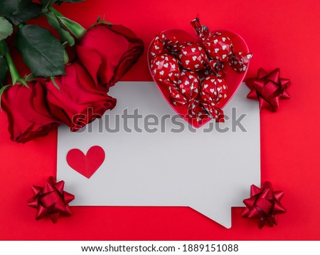 White sheet, three roses, ribbon, candy and a paper heart on a red background with place for text in the middle, top view close-up.