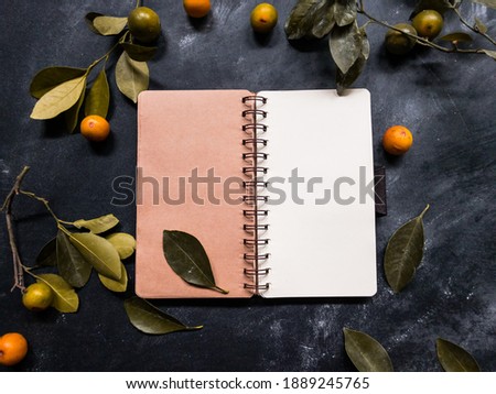 An open blank notebook surrounded by leaves and calamansi or kalamansi lime on black wooden table. Space for text. Selective focus. Copy space