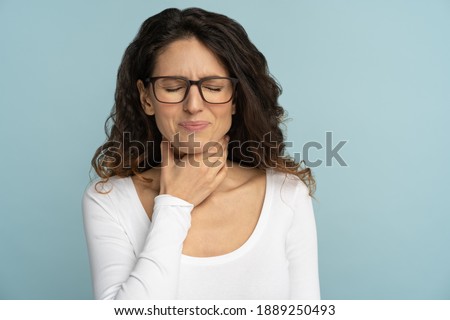 Sick woman having sore throat, tonsillitis, feeling sick, suffering from painful swallowing, angina, strong pain in throat, loss of voice, holding hand on her neck, isolated on studio blue background. Royalty-Free Stock Photo #1889250493