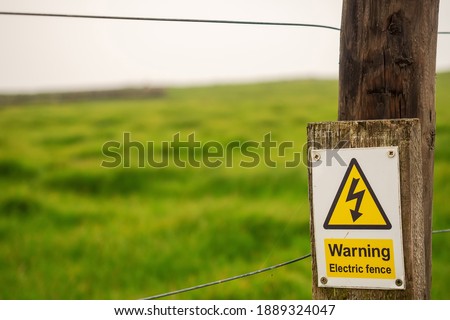 Warning electric fence sign on a wooden post. Green grass field out of focus, Agriculture industry concept