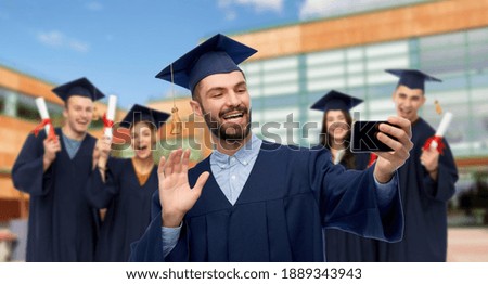 education, graduation and people concept - happy smiling male graduate student in mortar board and bachelor gown with smartphone having video call or taking selfie over school background