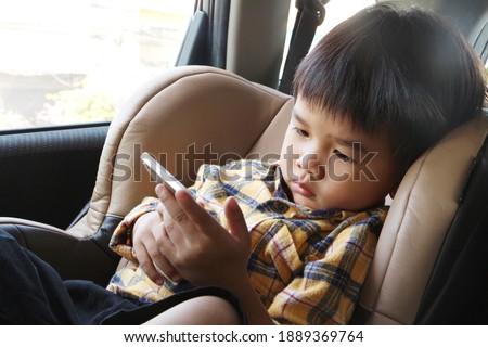 Asian boy looking at smartphone screen. Child sitting on car seat and watch video cartoon on mobile phone. Kid holding cellphone in his hand.
