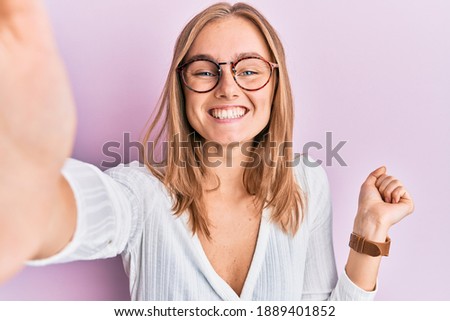 Beautiful blonde woman taking a selfie photo wearing glasses screaming proud, celebrating victory and success very excited with raised arm 