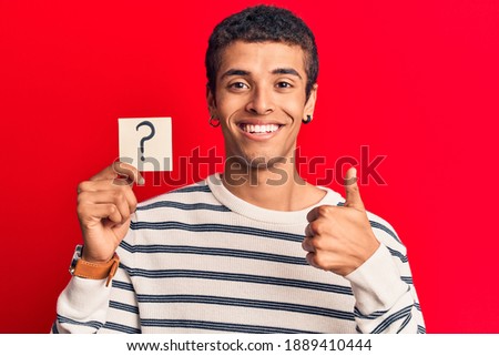Young african amercian man holding question mark reminder smiling happy and positive, thumb up doing excellent and approval sign 