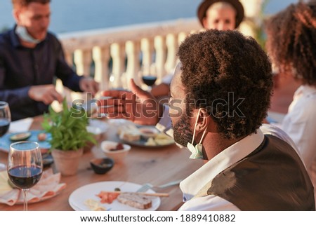Multiracial people enjoy dinner together outdoor on patio while wearing surgical face masks under chin - Coronavirus lifestyle and social distance concept