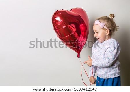 Little girl holding a heart air balloon and looks at it on a light background. Concept for Valentine's Day, birthday. Banner.