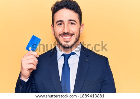 Young hispanic man wearing suit holding credit card looking positive and happy standing and smiling with a confident smile showing teeth 
