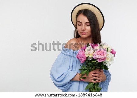 Teenage girl, happy looking woman with brunette long hair. Wearing a hat and blue dress. Holding a bouquet of beautiful flowers. Watching to the left lower corner at copy space over white background