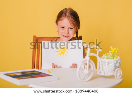 Little girl draws on a white sheet of paper. A creative child shows the sun hatch. Studio photo on a yellow background.