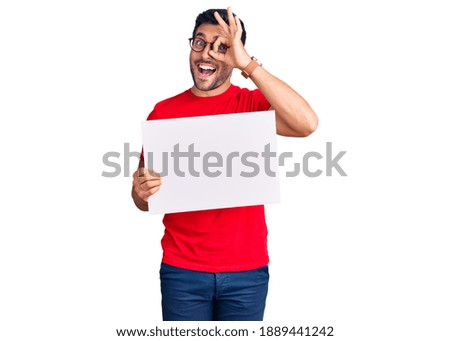 Young hispanic man holding blank empty banner smiling happy doing ok sign with hand on eye looking through fingers 