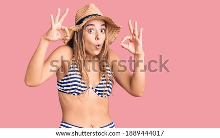 Young beautiful blonde woman wearing bikini and hat looking surprised and shocked doing ok approval symbol with fingers. crazy expression 