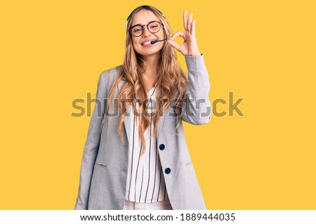 Young beautiful blonde woman wearing call center agent headset smiling positive doing ok sign with hand and fingers. successful expression. 