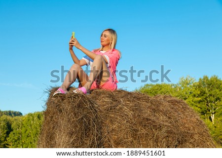 Blonde woman in bright pink suit and white T-shirt sits on haystack against background of mountains, trees and round rolls of hay with phone and takes selfie with hand outstretched. Summer vacation