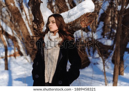 Girl in the black coat and white scarf at the background of the trees covered with snow. Sunny winter day. High quality photo