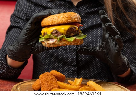 black gloves are holding a burger, nuggets and fries are on a wooden plate, fast food, a table in a bar, a close-up of a cheeseburger
