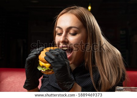 a young girl is biting a burger, there is beer on the table, a satisfied blonde face, a black shirt and gloves. food photo.