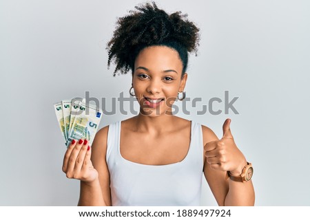 Young african american girl holding 5 euro banknotes smiling happy and positive, thumb up doing excellent and approval sign 