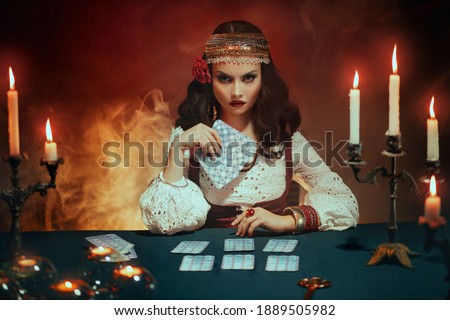 Fantasy beautiful girl in image of gypsy witch sits at table in dark gothic room. Art Red costume. Fortune teller magical woman reading future on tarot cards. Ritual candles burning, seance, smoke. Royalty-Free Stock Photo #1889505982