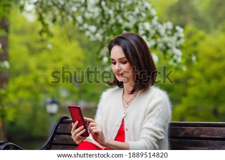 A model poses with a smile in a Park, sitting on an outdoor bench and talking on a smartphone in her hands. Portrait of a pretty brunette beauty girl with great makeup.