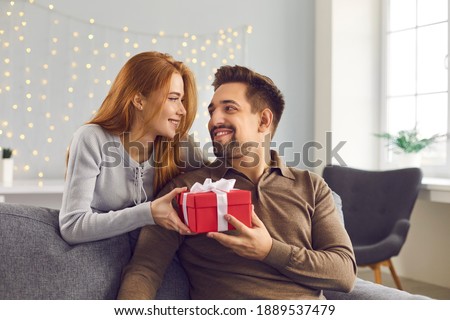 Beautiful young couple at home. Romantic woman stands behind a man sitting on a sofa and hands him a holiday present. Concept of gifts for birthdays, Christmas, Valentine's Day and Men's Day.