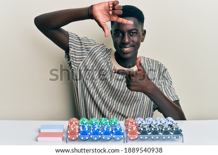 Young african american man sitting on the table with poker chips and cards smiling making frame with hands and fingers with happy face. creativity and photography concept. 