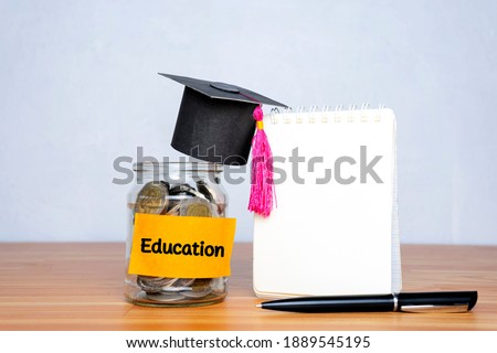 Graduation hat and glass jar with money, pen and a blank notebook for education on wooden table Royalty-Free Stock Photo #1889545195