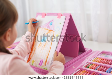 Child girl draws with colored pencils, masking illustration of her house, using multicolored felt tipped pens, big drawing set for children with many colored pencils and wax crayons.