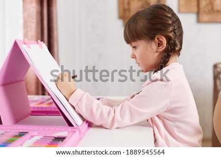 Profile portrait of drawing adorable talented female kid wearing casual shirt, making illustration with color pencils ar felt-tip pens, cute child sitting at desk in her room.