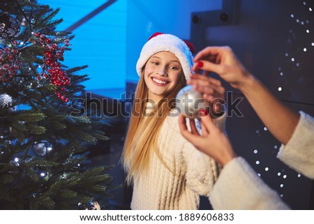 Happy girl is trimming New Year tree while her mother is helping her by giving glass balls