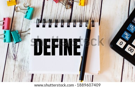 On a light wooden table lies a calculator, multi-colored paper clips and a notebook with a pen and the word DEFINE