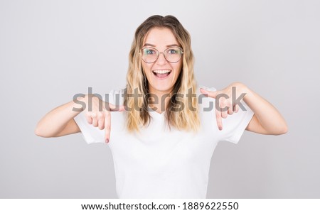 Photo of surprised blonde woman points on floor, has shocked facial expression, wears white t shirt against white background. Wow concept.