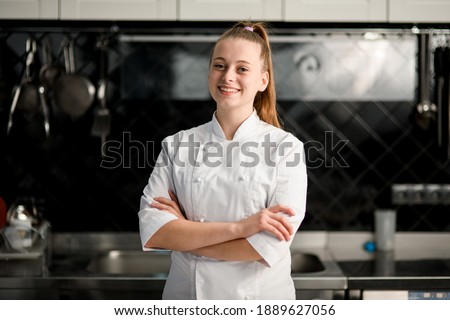 young beautiful smiling woman chef with arms crossed wearing in white suit at kitchen. Blurred background Royalty-Free Stock Photo #1889627056