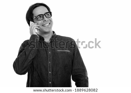 Studio shot of young happy Persian man smiling and talking on mobile phone while thinking