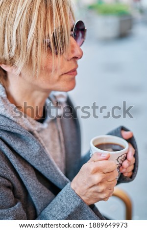 A Vertical photo of Caucasian blonde woman wearing sunglasses sitting in a bar terrace with coat having a cup of coffee