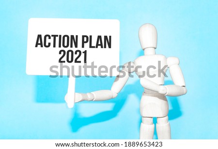 The wooden man holds a white sign with the text ACTION PLAN 2021 in his hands. The content of the lettering has implications for business concept and marketing.