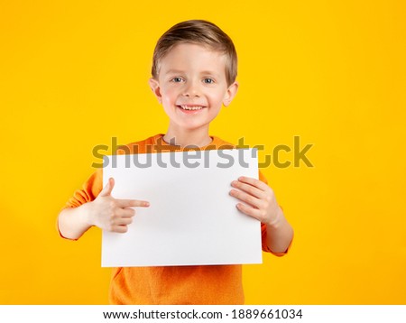 A beautiful European boy holds a clean white sheet of paper, points his finger at the place for text, advertising.