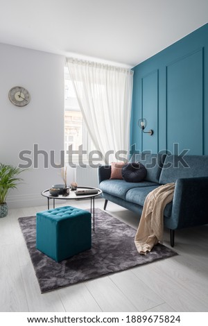 Stylish living room with teal blue wall with decorative molding, modern sofa, black coffee table and square, quilted ottoman Royalty-Free Stock Photo #1889675824