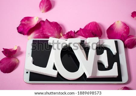 White wooden letters the word "love" on a smartphone In the pink background With rose petals all around Valentine concept. Valentine's Day background.