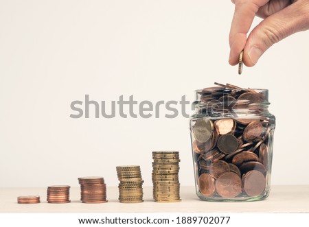 Stacked money coins in the form of a staircase to a jar filled with money coins, hand of a man puts a money coin in the jar, Concept for pension, reserve, savings or as a pension provision Royalty-Free Stock Photo #1889702077