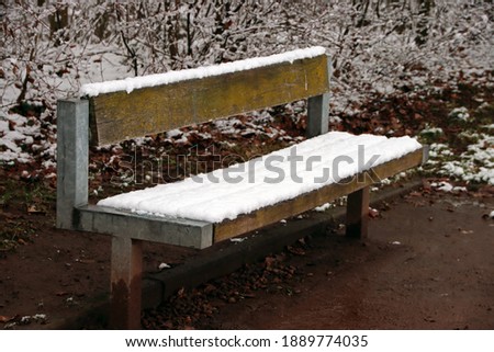 Snow-covered bench during winter in Germany, Kaiserslautern. Landscape and nature with snow and ice. Brown ground with dirt.