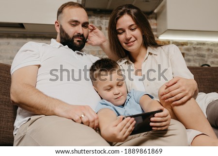 A dad, a son, and a mom are watching video on the sofa. A child is demonstrating a cartoon on the smartphone to mother and father in the evening at cozy home. 