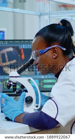 African woman scientist using microscope in modern equipped lab. Multiethnic team examining virus evolution using high tech for scientific research of treatment development against covid19. Royalty-Free Stock Photo #1889922067