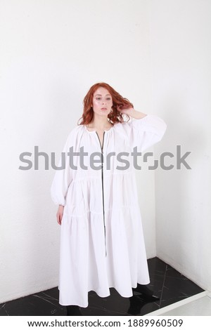 Catalog shooting of ginger model with pale skin in white oversize summer dress posing in spacious interior. Copy space. Model tests, changing different poses. Concept of new spring trends in fashion