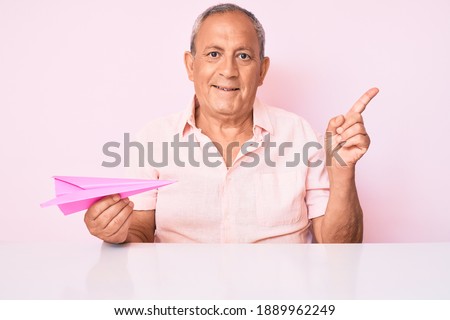 Senior handsome man with gray hair holding paper airplane smiling happy pointing with hand and finger to the side 