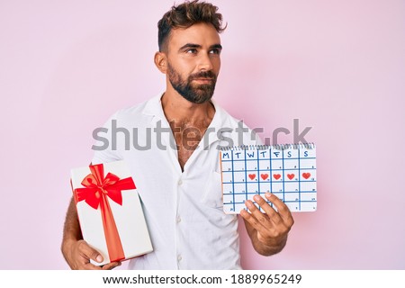 Young hispanic man holding heart calendar and paper airplane doing ok sign with fingers, smiling friendly gesturing excellent symbol 
