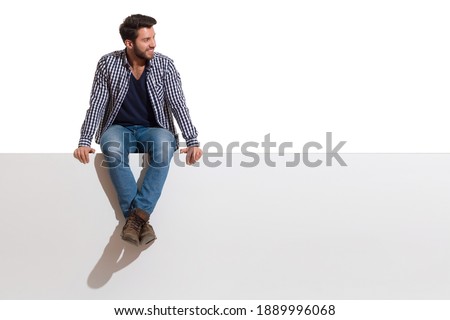 Handsome casual young man is sitting on a top of white banner with legs crossed, smiling and looking at the side. Full length studio shot isolated on white. Royalty-Free Stock Photo #1889996068