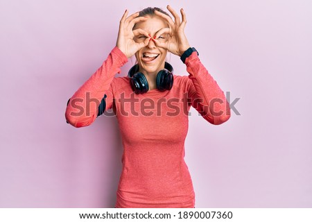 Beautiful caucasian woman wearing sportswear and arm band doing ok gesture like binoculars sticking tongue out, eyes looking through fingers. crazy expression. 
