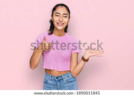 Hispanic teenager girl with dental braces wearing casual clothes showing palm hand and doing ok gesture with thumbs up, smiling happy and cheerful 