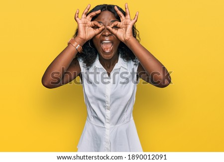 Beautiful african young woman wearing casual clothes doing ok gesture like binoculars sticking tongue out, eyes looking through fingers. crazy expression. 