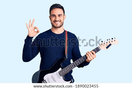 Young handsome man playing electric guitar doing ok sign with fingers, smiling friendly gesturing excellent symbol 